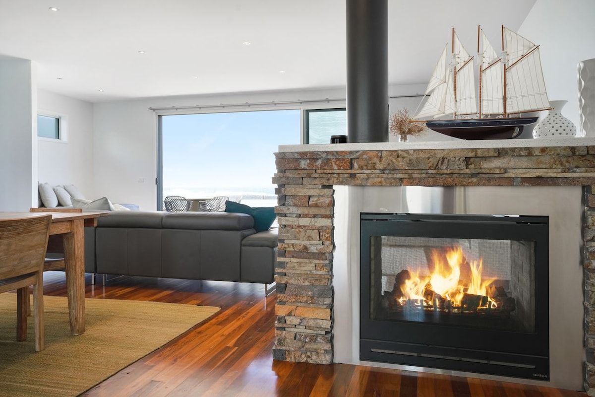 A sleek living area features a modern stone fireplace, which radiates warmth. A large window offers views of the ocean, while a comfortable sectional sofa is positioned nearby. A wooden dining table adds a natural element, and decorative items provide subtle accents.