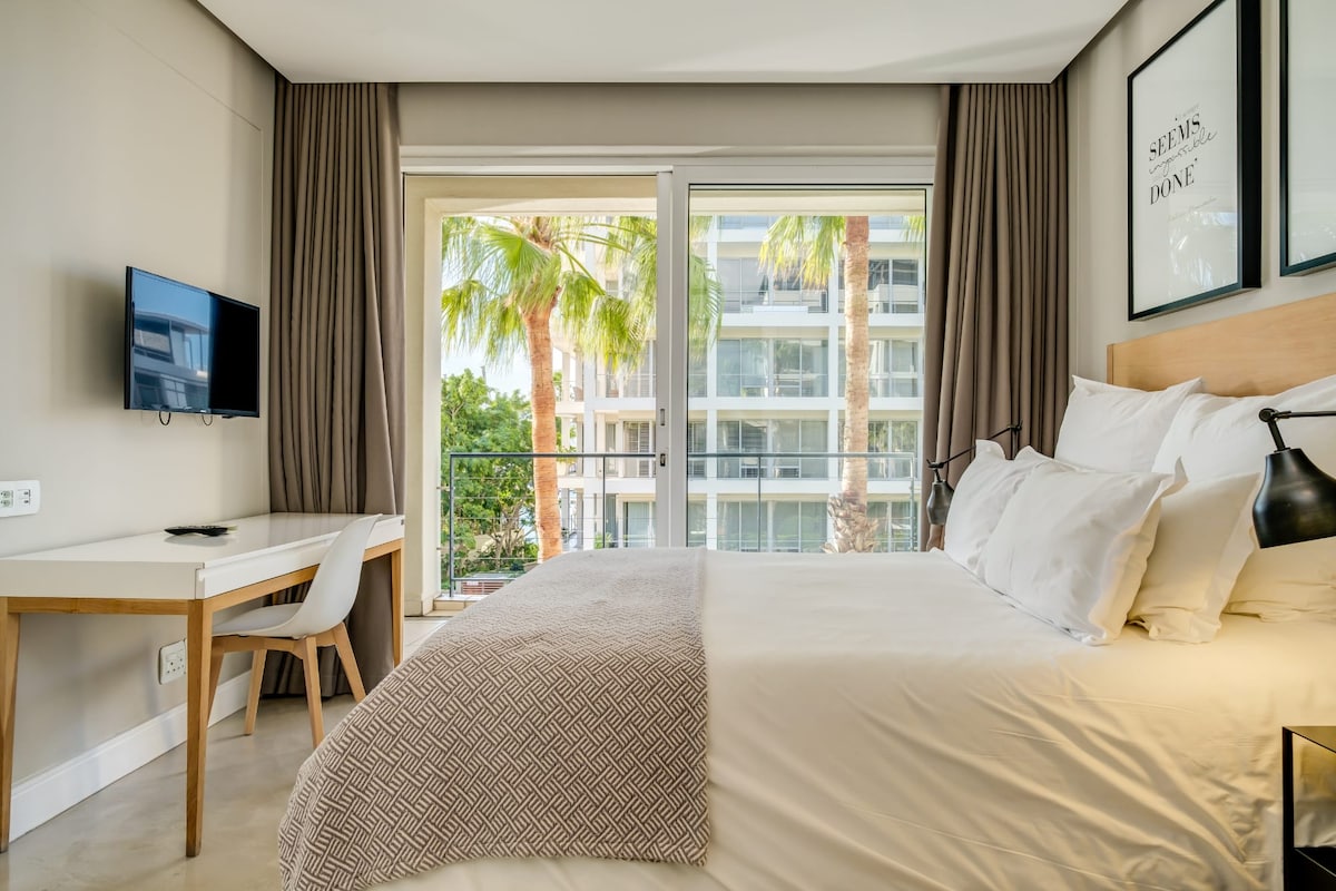 A modern sleeping area features a spacious bed against a light-colored wall. Large windows allow natural light and offer views of palm trees outside. A small desk and a wall-mounted television are present, enhancing the functional layout of the room.