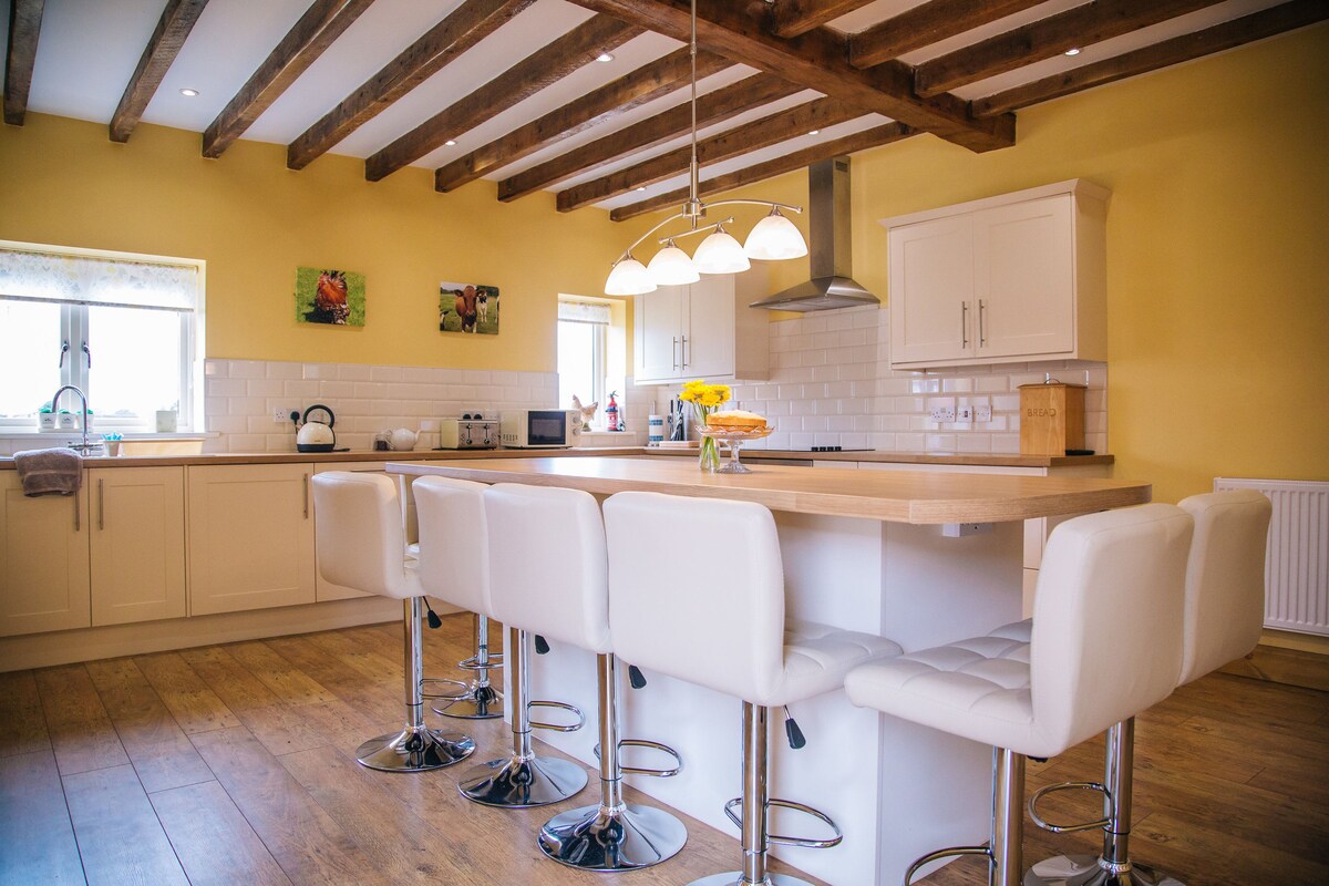 A spacious kitchen features modern appliances and a large island with seating for six. Warm yellow walls complement the wooden ceiling beams. Large windows allow natural light to enhance the inviting atmosphere. Decorative art can be seen on the walls.