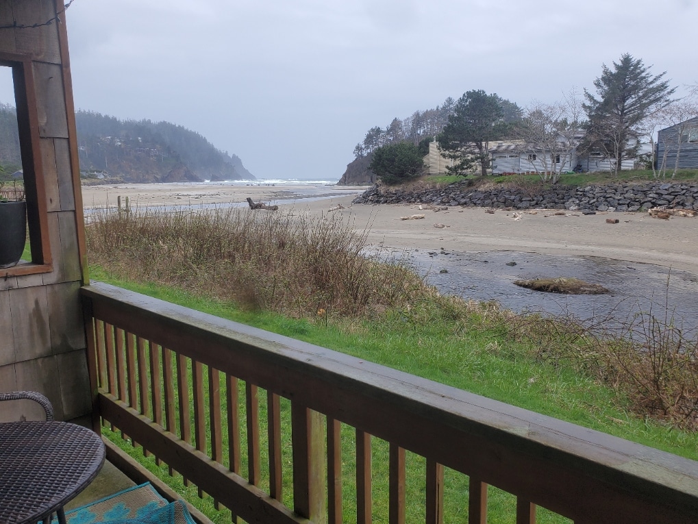A view from a wooden patio railing showcases a serene coastal landscape. Waves gently roll onto the sandy beach, with rocky outcrops visible along the shoreline. A backdrop of rolling hills and sparse vegetation frames the scene, creating a calm, natural atmosphere.