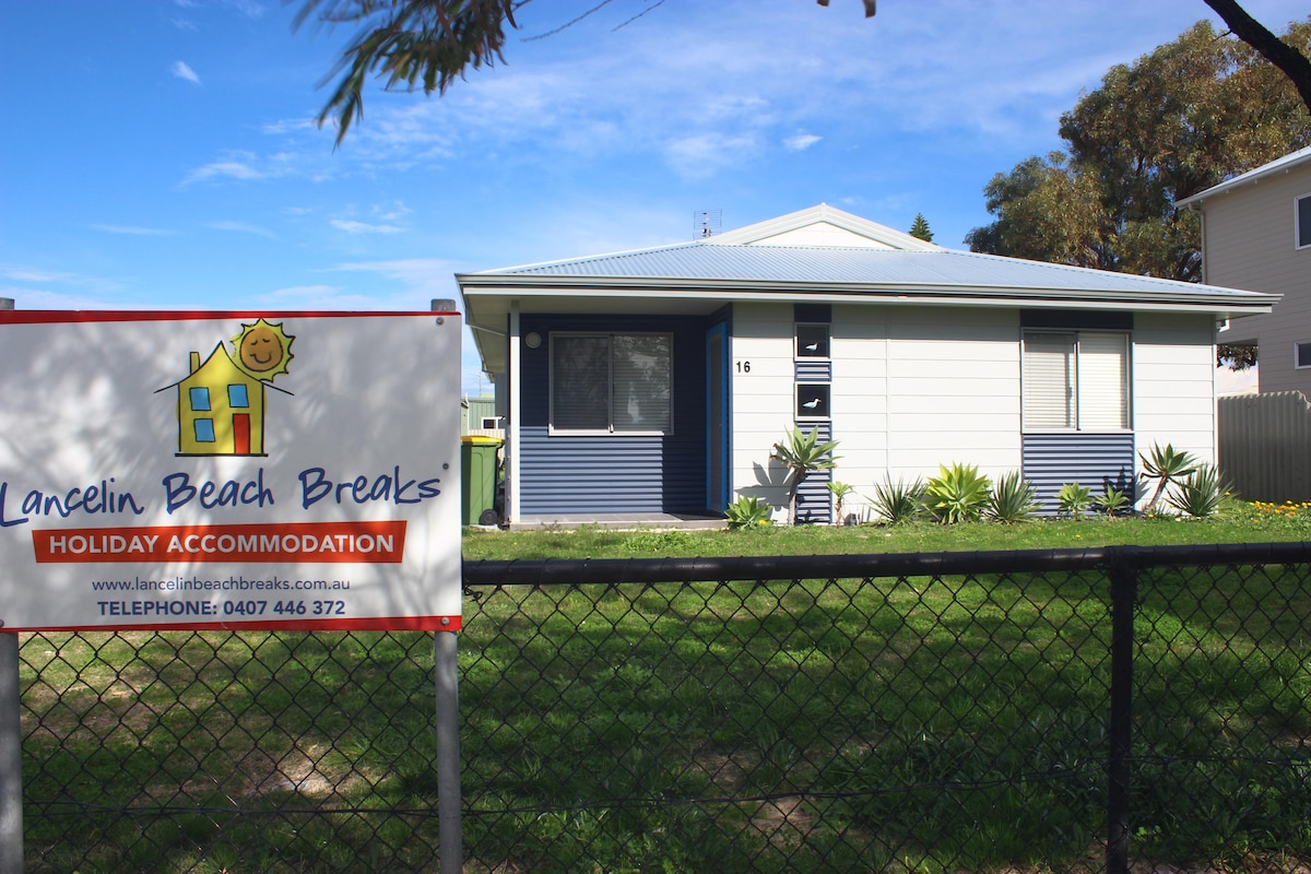 A single-story beach cottage is set behind a chain-link fence, showcasing a neat exterior with contrasting colors. The front yard features green grass and low-maintenance landscaping. A sign highlighting the holiday accommodation is prominently displayed in front of the cottage.