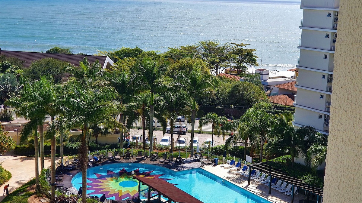 A clear view of the inviting pool area is presented, surrounded by palm trees and colorful lounge chairs. The ocean is visible in the background, reflecting sunlight, while the vibrant pool design adds a touch of color to the serene setting.