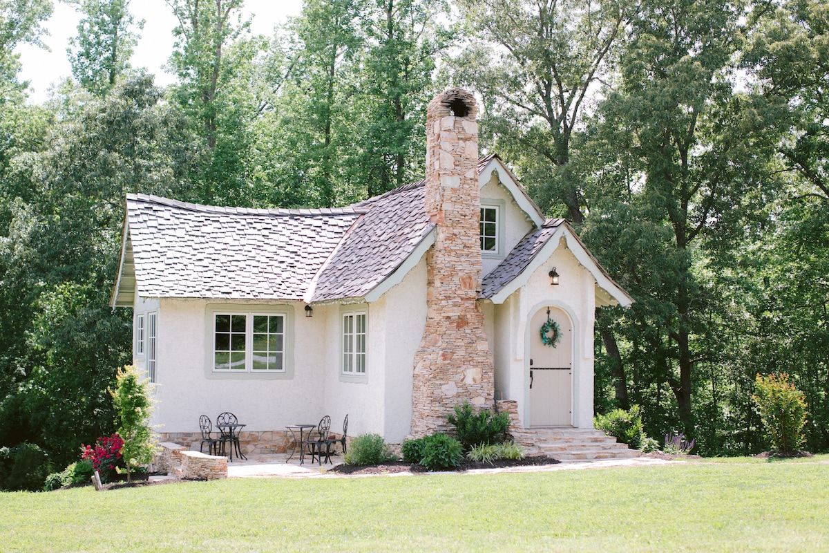 A charming cottage exterior is showcased with a stone chimney and a welcoming front door adorned with a wreath. Surrounded by lush greenery, a small outdoor seating area with two chairs is nestled beside vibrant flowerbeds.