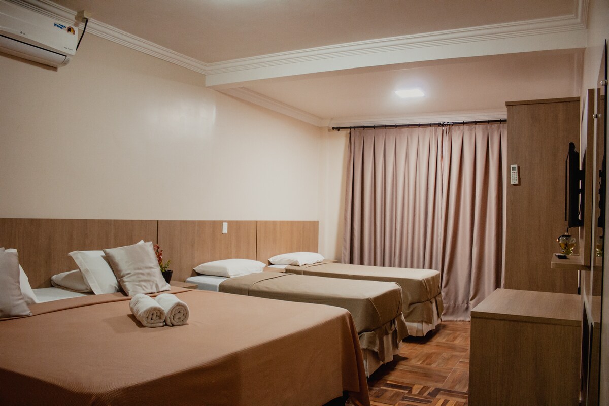 A spacious room is furnished with two beds and a single bed, adorned with neutral-toned linens. Soft lighting illuminates the area, complemented by curtains that frame the window. A wooden cabinet and a wall-mounted television are also present, enhancing convenience.