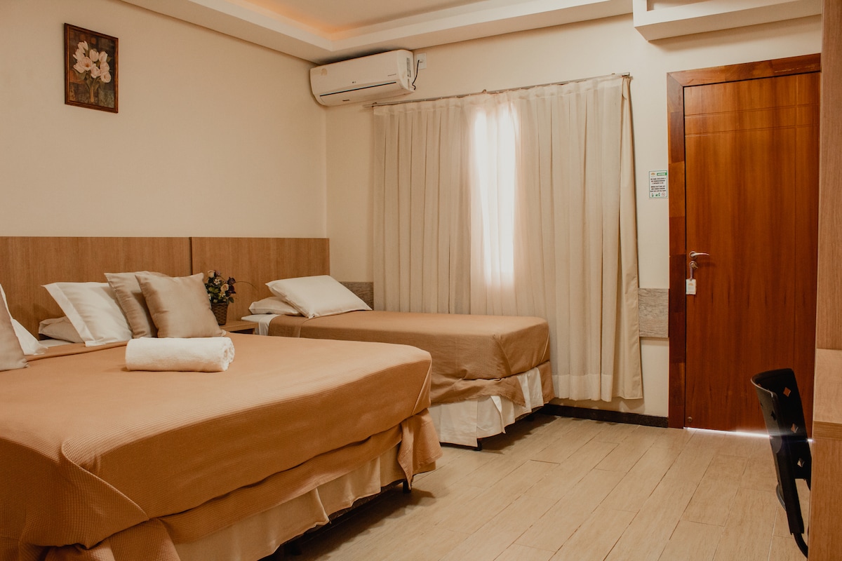 A spacious room is furnished with two beds, featuring neutral-toned bedding. A light-colored curtain gently diffuses natural light from a window. An air conditioning unit is mounted on the wall, and a piece of wall art adds subtle decoration above the beds.