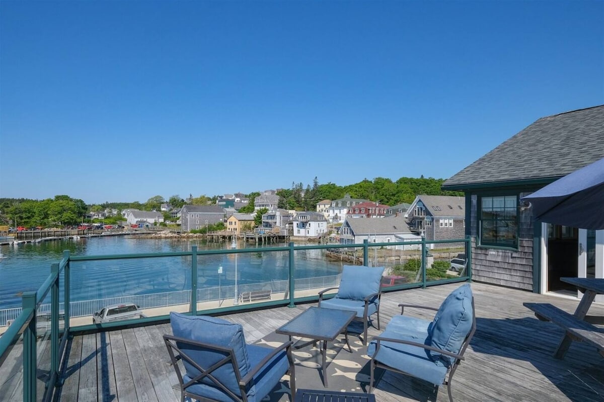 A spacious deck features comfortable seating arrangements, including two lounge chairs and a table, overlooking the tranquil Stonington thoroughfare. The bright blue sky contrasts with the quaint village, where colorful buildings are visible along the shoreline, enhancing the scenic waterfront view.