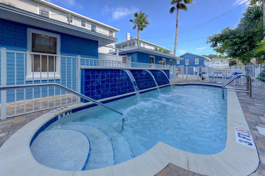 A heated pool is presented, featuring three waterfalls gently flowing into the water. Paver pathways and a safety railing outline the pool area, which is surrounded by palm trees and blue cottage exteriors, creating a relaxing outdoor atmosphere.