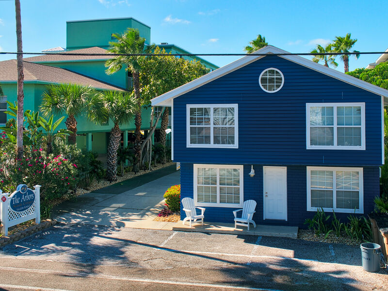 The two-story cottage is showcased with a blue exterior and white trim. Two white adirondack chairs are positioned on a small porch, surrounded by tropical landscaping. The clear sky and vibrant greenery create a welcoming atmosphere. A neighboring building is visible in the background.