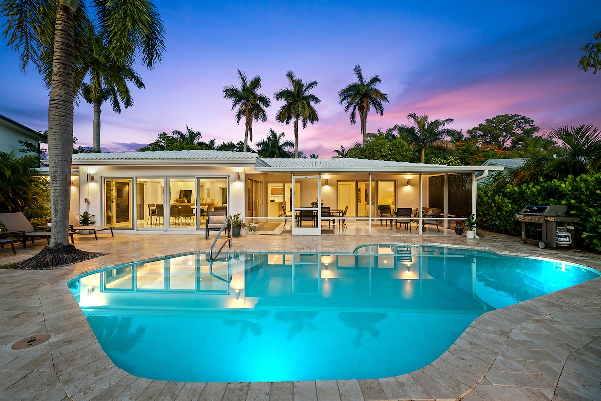 A serene outdoor space features a heated pool reflecting the evening sky, surrounded by palm trees. Comfortable seating areas are visible, along with a grill on the side. Large glass doors provide an unobstructed view of the interior spaces, enhancing the connection with nature.