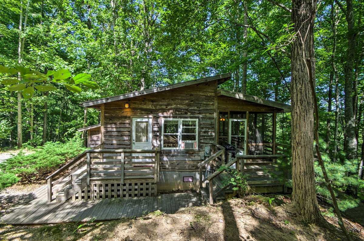 A rustic cabin is nestled among lush greenery, featuring a wooden exterior and a welcoming front porch. Large windows invite natural light, and the expansive deck provides ample outdoor space for relaxation surrounded by trees.