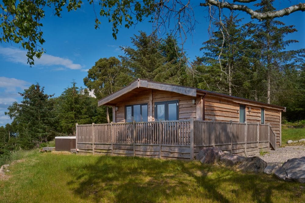 The lodge is positioned among trees, showcasing wooden exterior and large windows. A spacious deck extends from the front, surrounded by grassy areas and rocky features. A hot tub is visible on the side, contributing to the peaceful atmosphere of the rural setting.