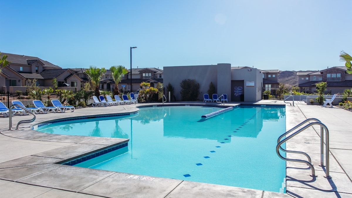 A swimming pool is surrounded by lounge chairs and palm trees. The clear, blue water reflects the sunlight, creating a serene atmosphere. Adjacent buildings are visible in the background, contributing to the overall community setting. Pathways and safety rails enhance accessibility.