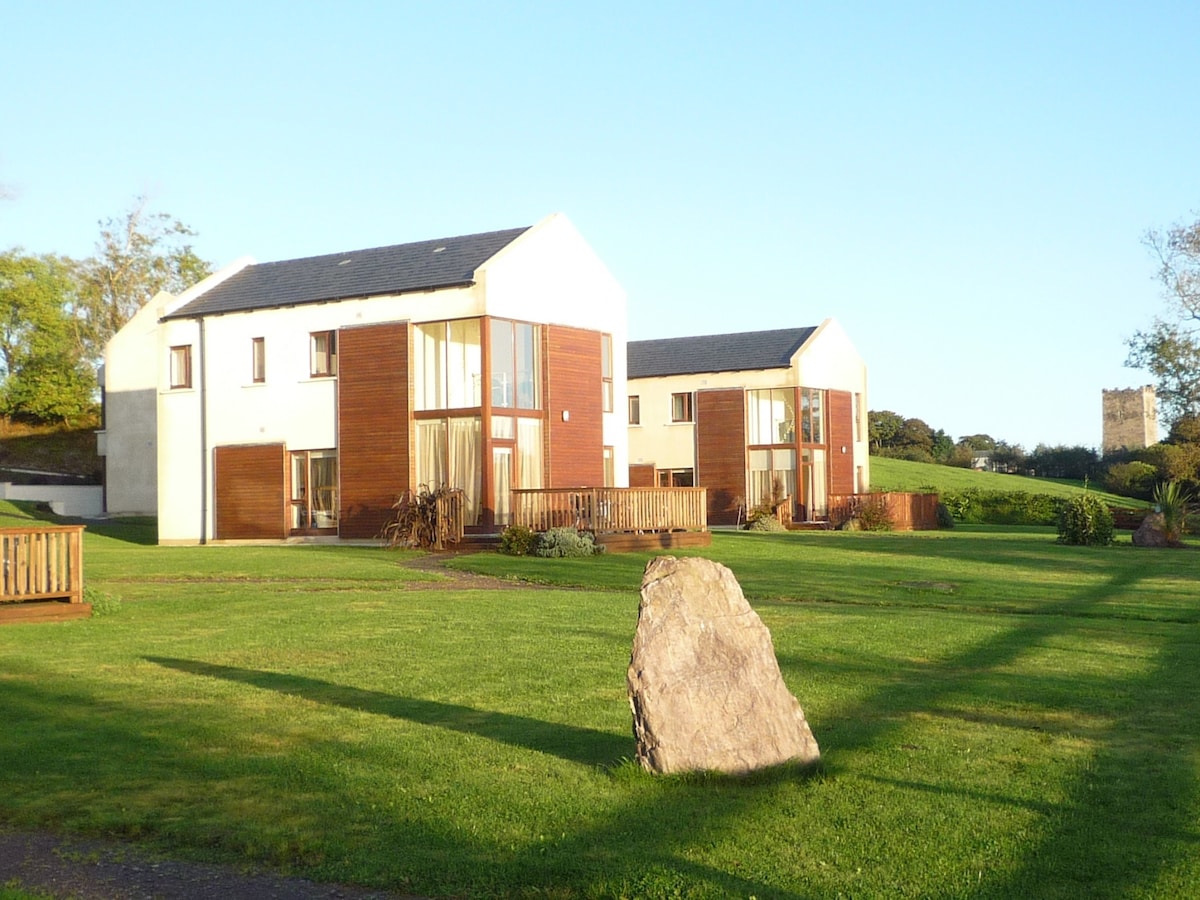 Two large, modern holiday homes are set among well-manicured lawns. The buildings feature a combination of white walls and wooden accents. A sizable rock is positioned in the foreground, surrounded by lush greenery. A clear blue sky complements the tranquil setting.