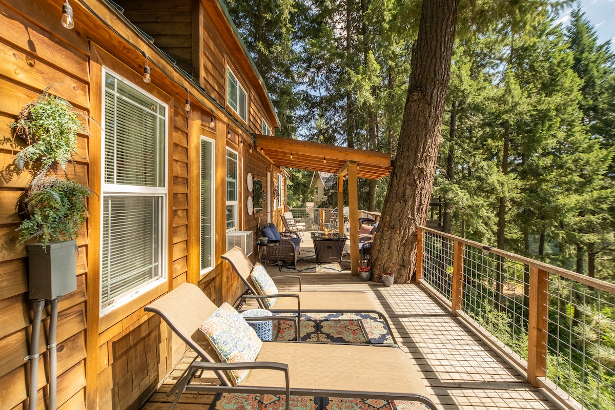 A spacious wraparound deck is highlighted with loungers and potted plants, offering serene views of the surrounding forest. The wood exterior of the cabin complements the natural beauty, while the inviting outdoor seating area is designed for relaxation in a tranquil setting.