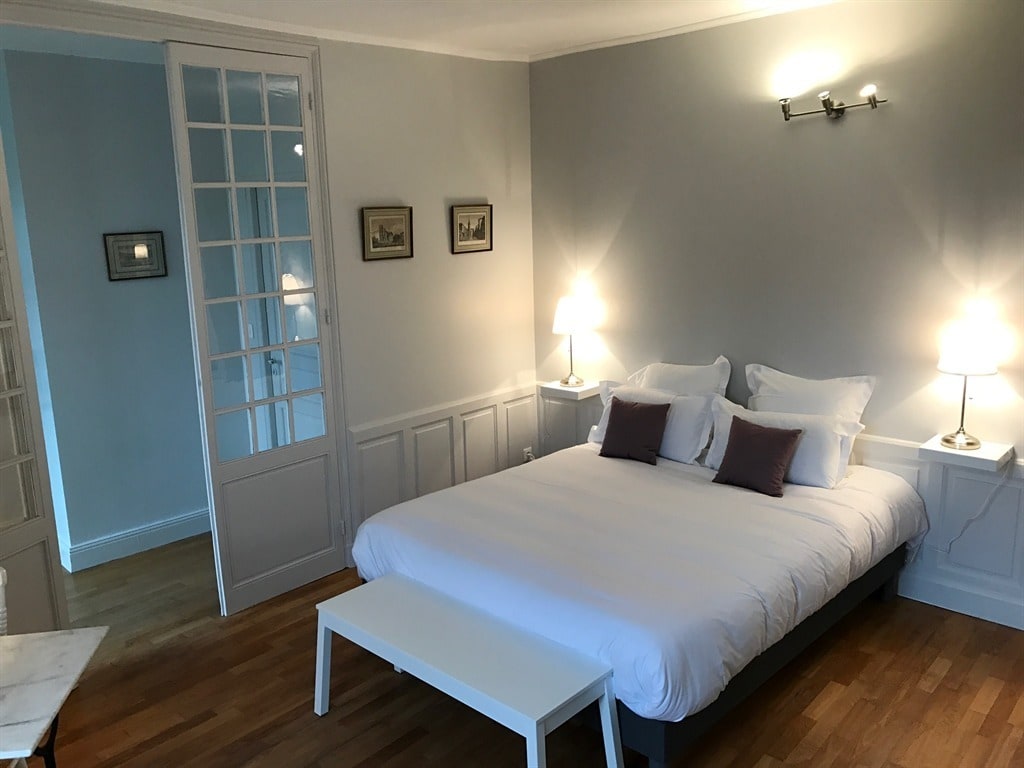 A well-lit bedroom features a spacious bed with white linens and two decorative pillows. Nightstands with lamps are positioned on either side of the bed. Light hardwood flooring complements the soft gray walls, and glass doors lead to another area of the apartment.