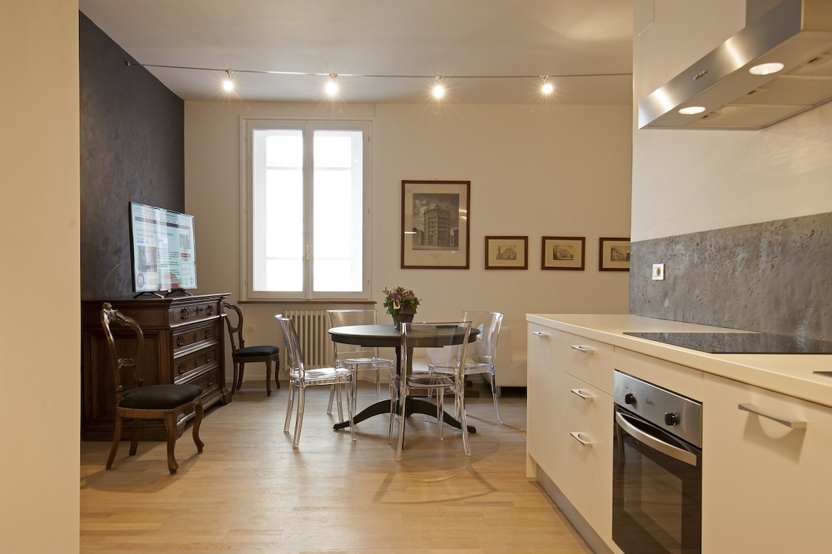 An open kitchen and dining area features a round table surrounded by clear chairs. A wooden sideboard adds character, while artwork adorns the walls. Natural light enters through large windows, creating a bright and functional space for dining and relaxing.