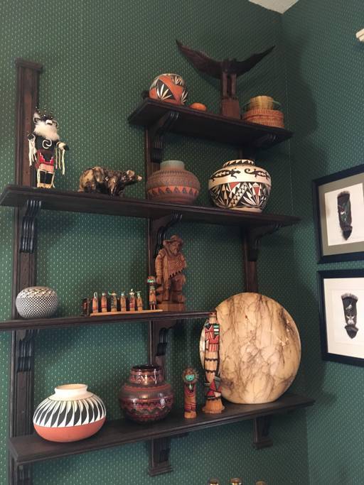 Shelves are adorned with a variety of decorative artifacts, including hand-crafted pottery, small figurines, and a wooden sculpture. The items display a range of colors and intricate patterns, all set against a rich green wall. Two framed artworks are visible alongside the shelves.