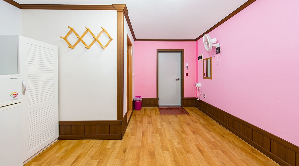 A bright interior space features soft pink walls, wooden trim, and a warm wooden floor. A small door leads to the outside, while a mirror and a wall-mounted fan are visible. Hat hooks are affixed to one wall, and a wastebasket is located nearby.