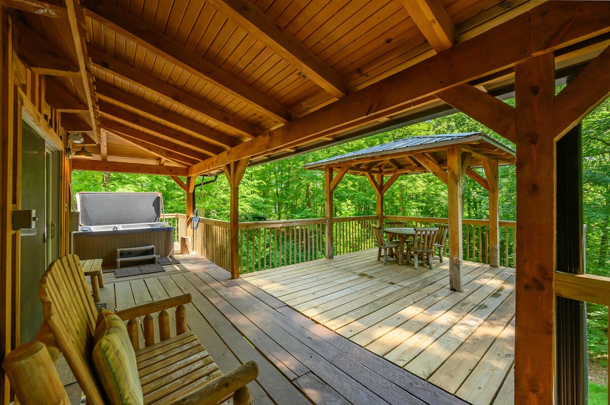 A spacious wooden deck is surrounded by trees, providing a natural retreat. The area features comfortable seating and a hot tub, with an open structure providing shelter and a view of the forest. The warm wooden tones create a welcoming atmosphere.