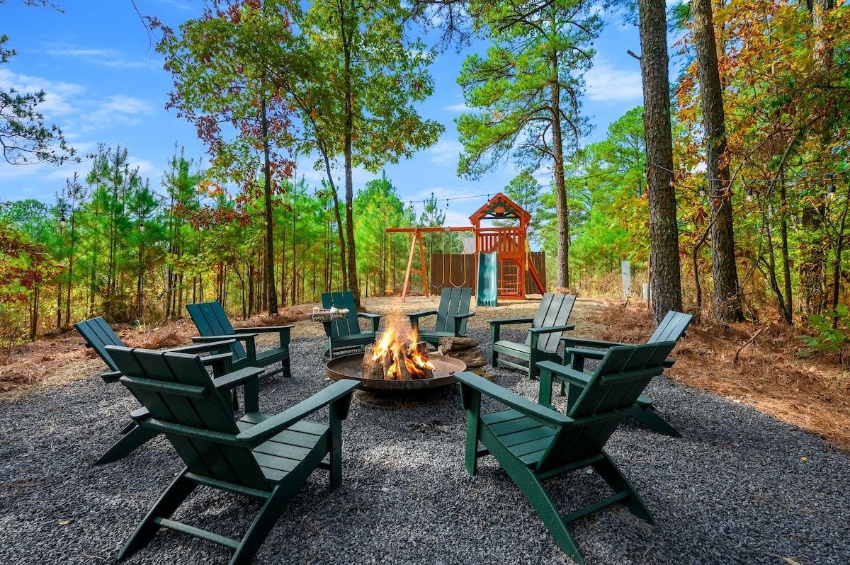 An outdoor fire pit area is surrounded by eight green Adirondack chairs, positioned on gravel. In the background, a children's playground features a playset and swings, framed by tall trees and a clear blue sky.