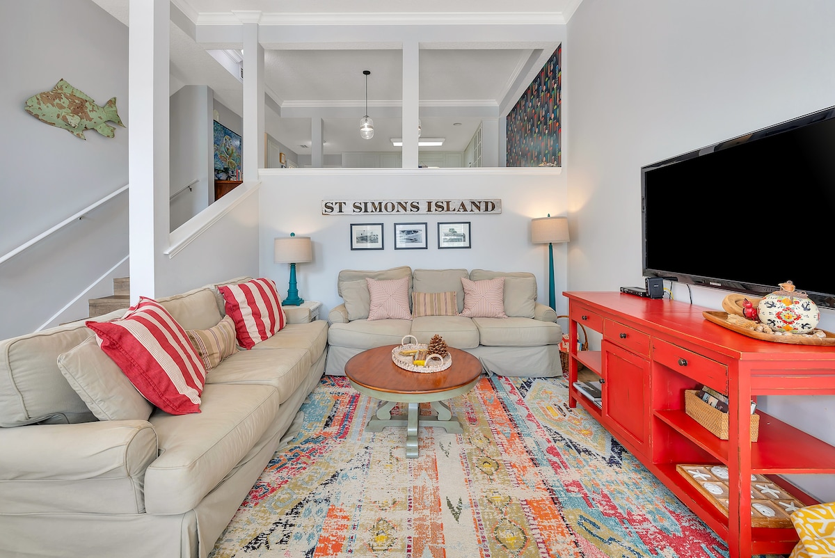 A comfortable living room is presented, featuring a light-colored sofa adorned with red and white striped pillows. A round coffee table sits on a vibrant area rug, with a colorful console table and television against the wall. Decorative lamps provide soft illumination.