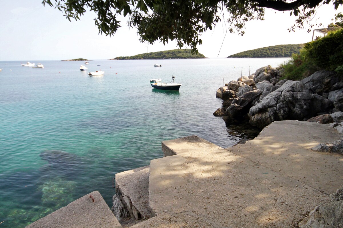 The image presents a serene coastal view featuring a concrete slab beach and clear turquoise waters. Several small boats are anchored nearby, with lush green islands visible in the background. The scene is shaded by overhanging branches, enhancing the tranquil atmosphere.