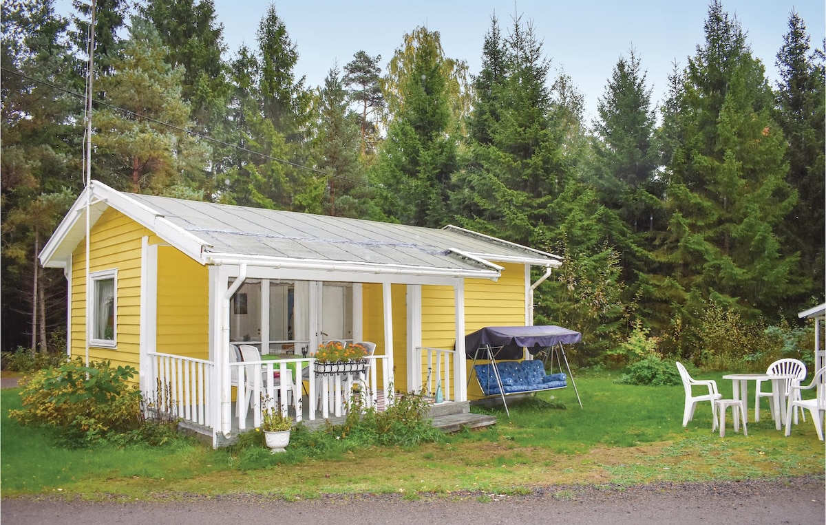 A simple cottage with a yellow exterior is surrounded by lush green trees. A covered porch features a seating area and a swing. The well-tended lawn includes a few outdoor chairs and a table, providing a welcoming space for relaxation.