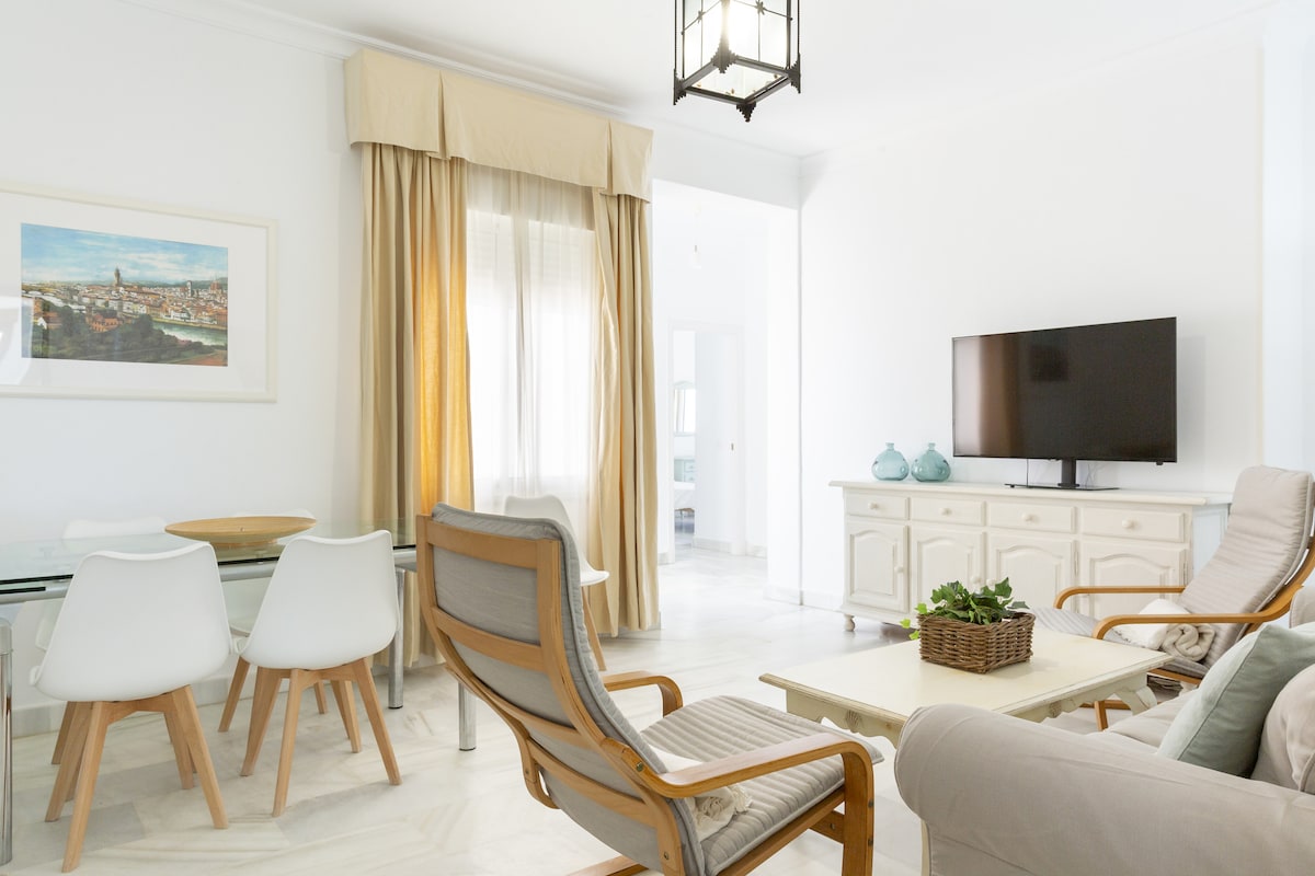 A bright and airy living space is featured, consisting of a comfortable seating area with various chairs and a small table. Natural light enters through the large window, contrasted by soft drapes. A television is positioned atop a light-colored sideboard, enhancing the open ambiance.