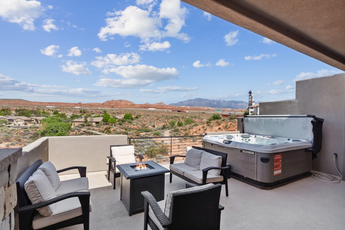 A spacious balcony features a private hot tub and a sleek fire pit surrounded by comfortable seating. Scenic views of red rock landscapes and blue skies are visible in the background, creating a peaceful outdoor atmosphere.