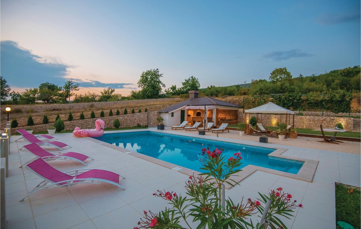 A spacious outdoor area features a large swimming pool surrounded by loungers and colorful greenery. A covered terrace with seating is nearby, alongside a charming summer kitchen. The backdrop includes lush trees and a picturesque sky at dusk, creating a serene atmosphere.