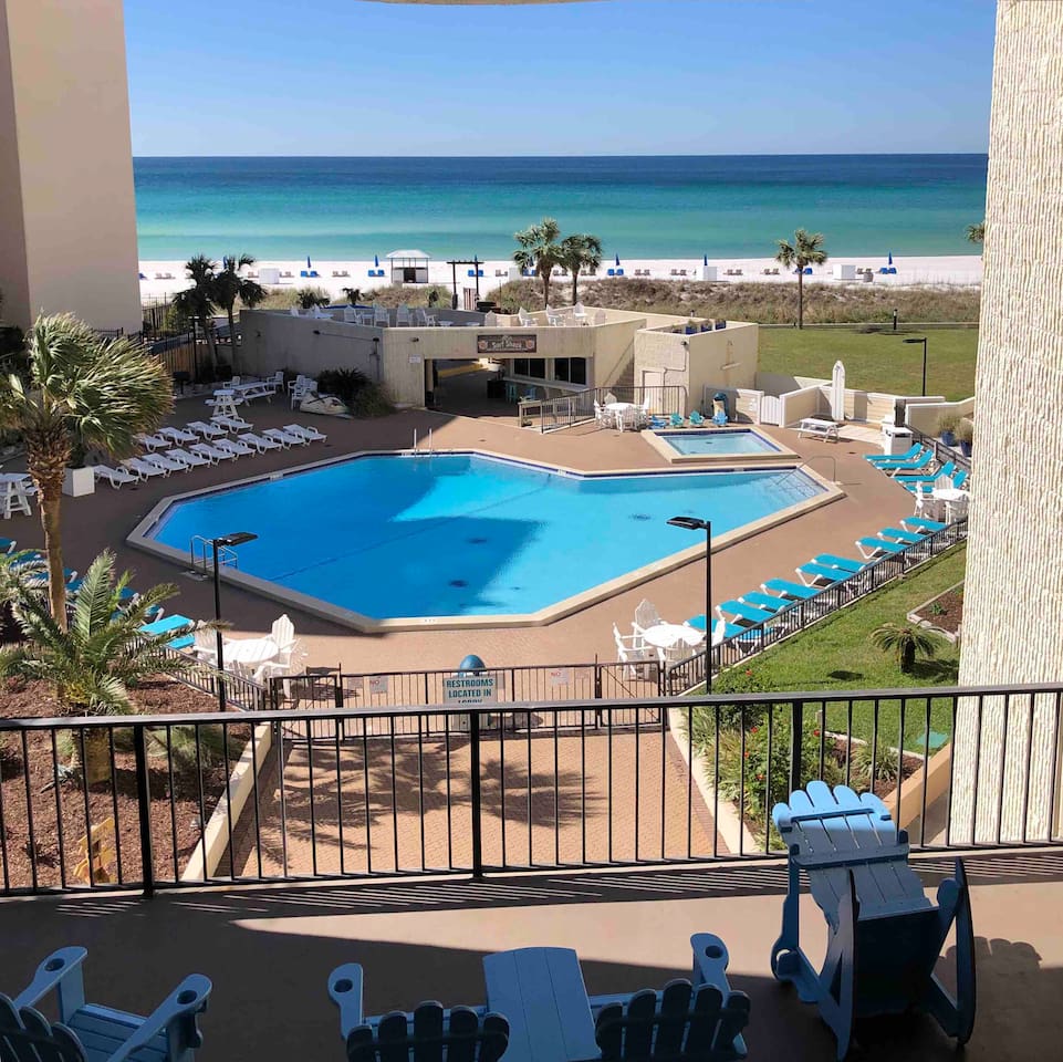 A spacious pool area is surrounded by lounge chairs, with a clear view of the sandy beach and ocean in the background. The pool's shape is octagonal, and a shaded seating area is visible nearby. Palm trees offer a touch of greenery.