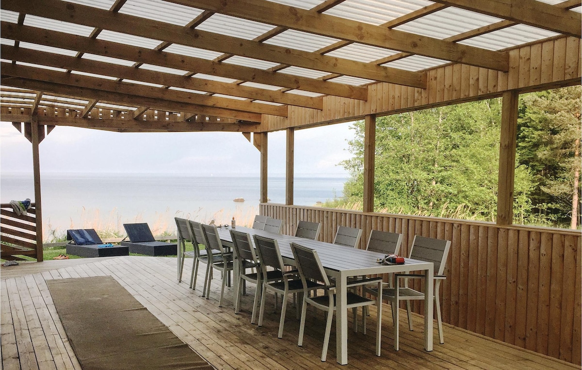 A spacious covered terrace features a long wooden table surrounded by multiple chairs. Large glass panels provide unobstructed views of the nearby lake and surrounding greenery, creating an inviting setting for gatherings. The floor is made of wooden planks, enhancing the natural ambiance.