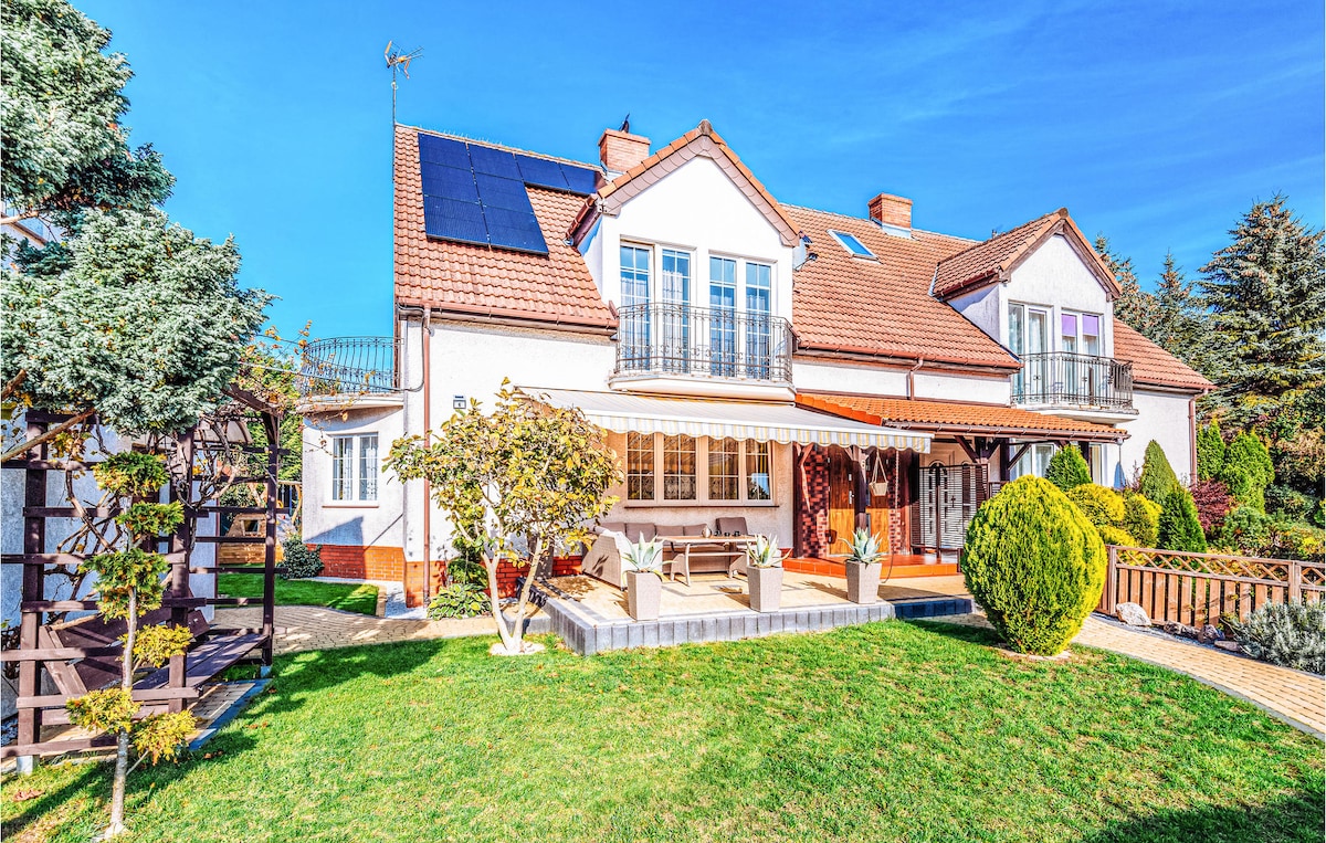 A semi-detached house is depicted, featuring a well-maintained exterior with a sloped roof and solar panels. Lush greenery surrounds the property, and a patio area is visible with seating. A wooden deck and a staircase lead to an upper level, showcasing outdoor spaces for relaxation.