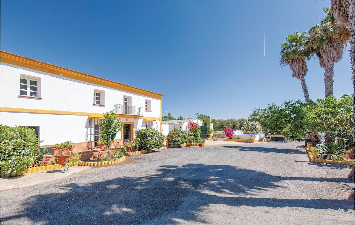 A spacious courtyard is framed by vibrant greenery and flowering plants, with a traditional two-story building featuring white walls and a yellow trim on one side. A clear blue sky enhances the inviting atmosphere of the outdoor area.