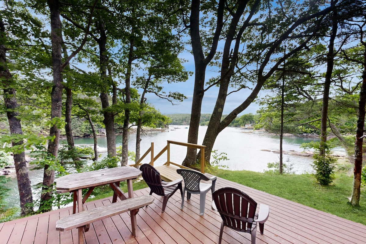 An outdoor deck is shown overlooking an ocean cove, surrounded by trees. A wooden picnic table and several adirondack chairs are positioned on the deck, inviting relaxation while enjoying scenic views of the water and natural landscape.