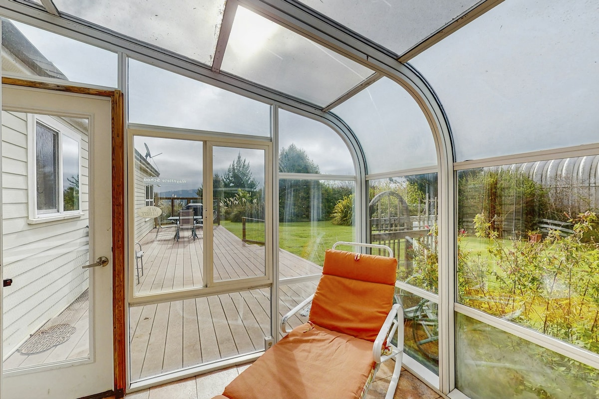 A sunroom is equipped with a lounge chair in a warm orange hue, allowing for relaxation while enjoying views of the surrounding garden and outdoor deck. Natural light filters through the clear walls and ceiling, enhancing the serene atmosphere.