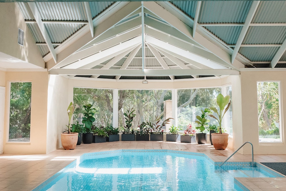 An indoor heated pool is framed by large glass windows, allowing natural light to fill the space. Elegant potted plants line the interior, and the ceiling is adorned with exposed beams, enhancing the open and airy atmosphere.