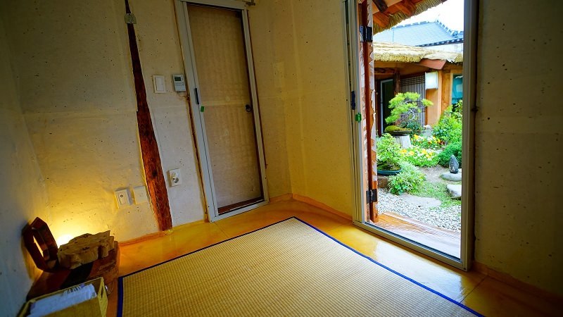 A serene interior space is presented with wooden doors leading to a vibrant garden area. Natural light fills the room, highlighting the tatami mat flooring. A small decorative lantern sits in one corner, contributing to the calm atmosphere created by the gentle colors of the walls.