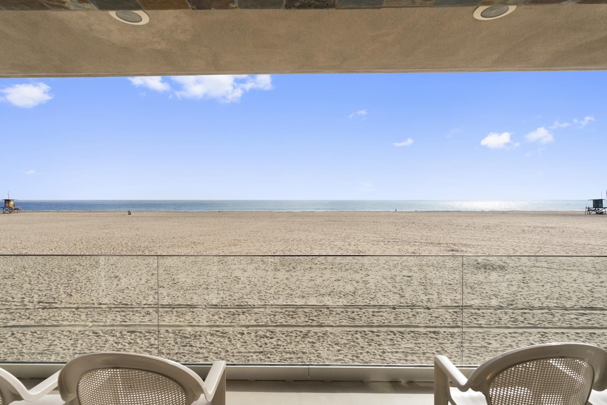 An expansive balcony view captures the sandy beach and the ocean beyond. The bright blue sky meets the water at the horizon, with distant figures enjoying the beach. Two lightweight chairs are positioned in the foreground, offering a spot to relax while enjoying the view.