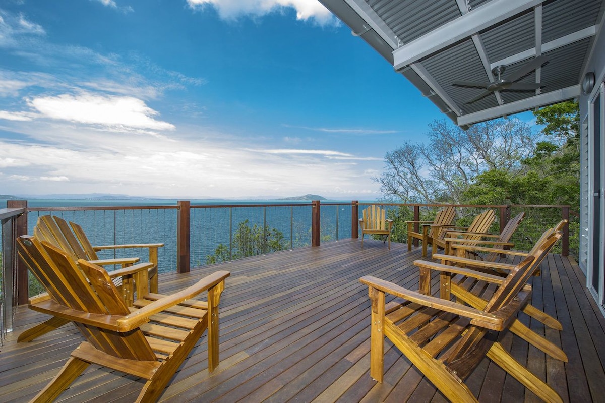 A spacious wooden deck offers stunning views of the Coral Sea, with several Adirondack chairs arranged for relaxation. The open sky presents a blend of soft clouds and blue tones, creating a serene outdoor space for enjoying the natural surroundings.