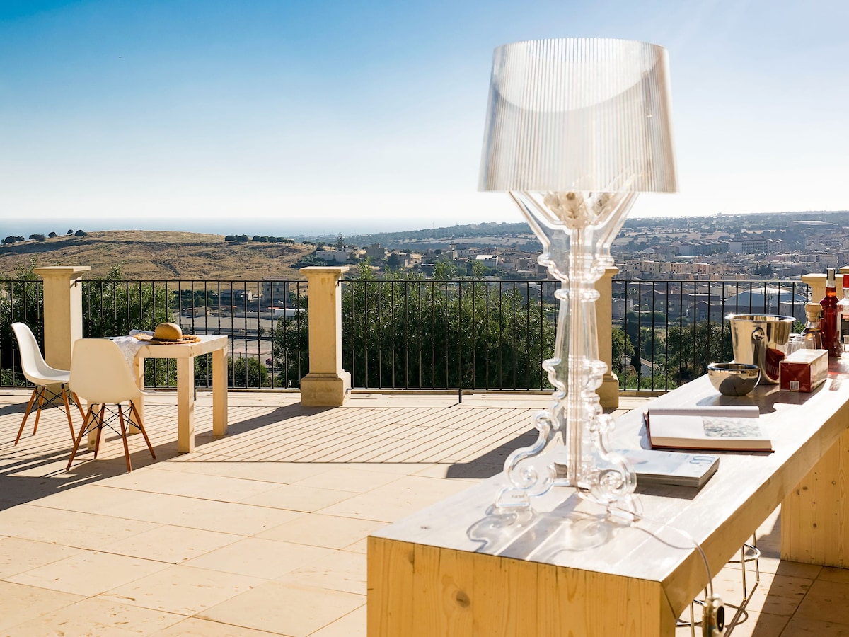 A spacious terrace features an elegant transparent lamp on a wooden tabletop, complemented by a dining table and chair set. Expansive views of the countryside and distant hills extend under a clear blue sky, providing a serene outdoor setting.