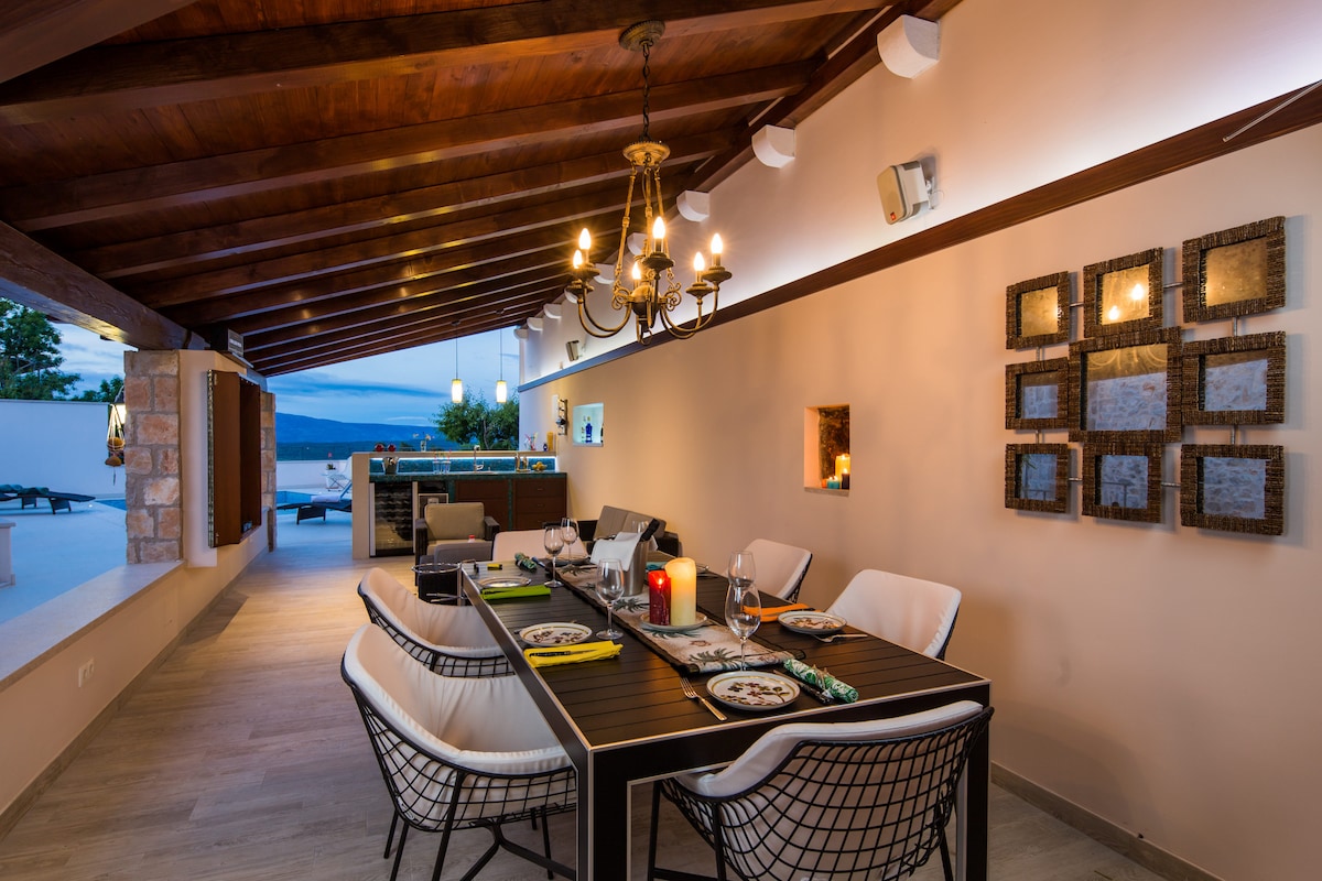 An outdoor dining area is showcased under a wooden beamed ceiling. A large table is set with dinnerware, surrounded by modern chairs. Soft lighting from a chandelier and wall sconces creates a warm ambiance, while views of the terrace and pool area can be seen in the background.