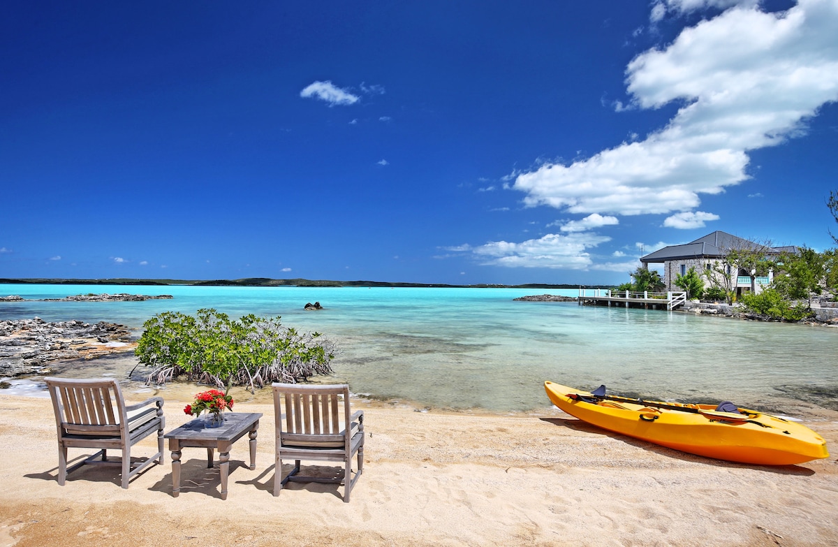 The image shows a sandy beach area leading to clear turquoise waters. Two wooden loungers are positioned on the shore beside a small table with flowers. A bright yellow kayak rests on the sand, while a dock and villa are visible in the background, framed by lush greenery.