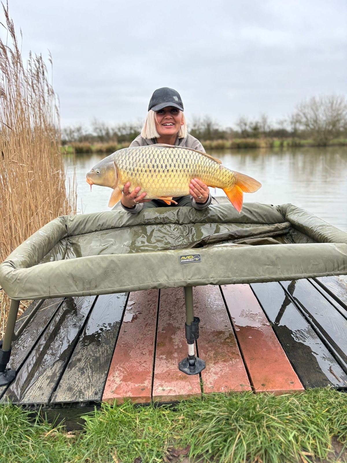 A person is seated on a fishing platform, holding a large fish with distinctive scales. Water surrounds the platform, while tall reeds frame the scene. The sky is overcast, suggesting a calm day by the lake.