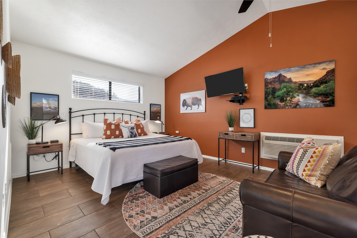 The room features a spacious bed with a patterned throw and decorative pillows. A mounted flat-screen TV is positioned on the wall. Natural light enters through the window, complemented by earthy tones and a patterned area rug on the tiled floor. Comfortable seating is provided by a leather sofa.
