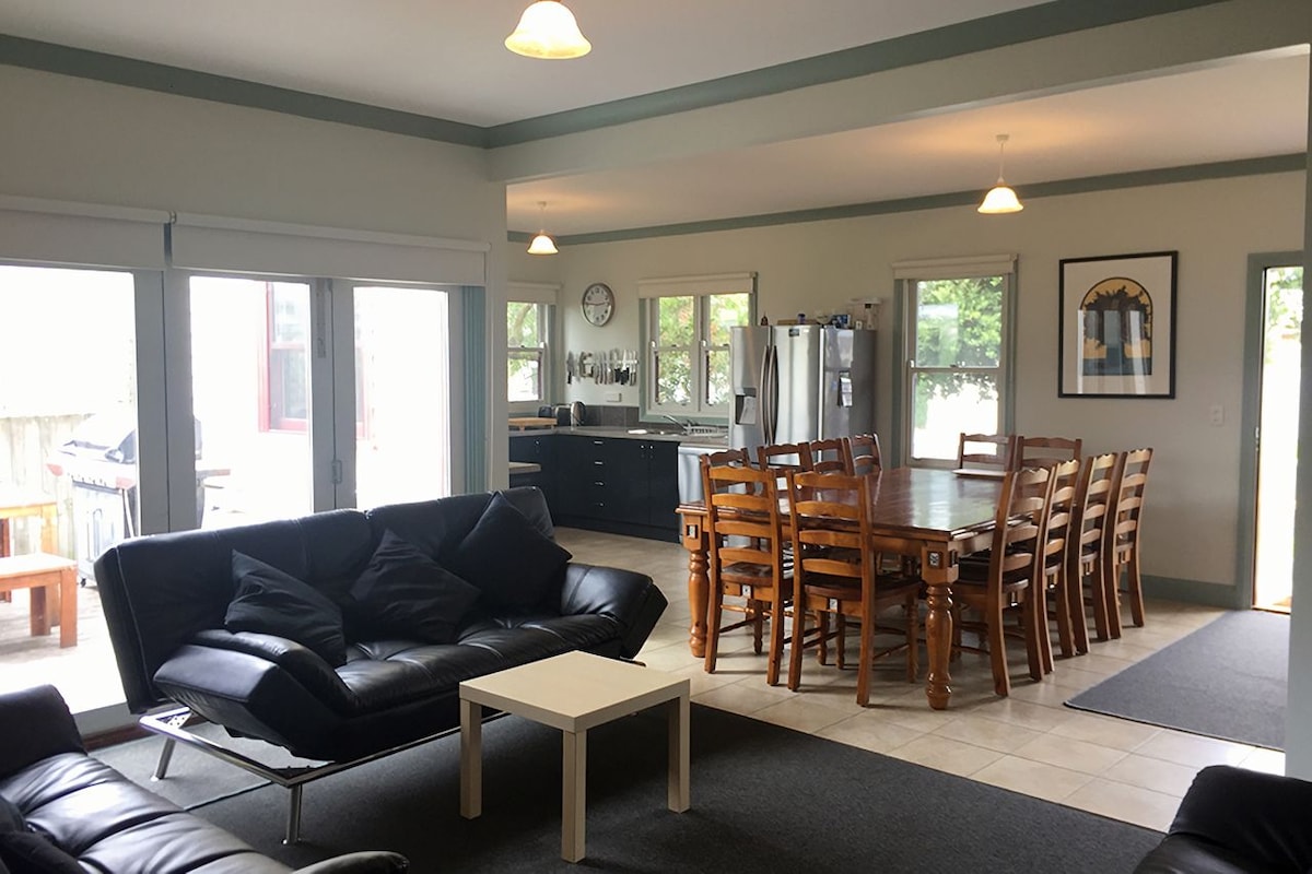 An open-plan living area features a dark leather sofa and a light coffee table, complemented by a large dining table that seats twelve. Abundant natural light is provided by multiple windows, and a well-equipped kitchen is visible in the background.