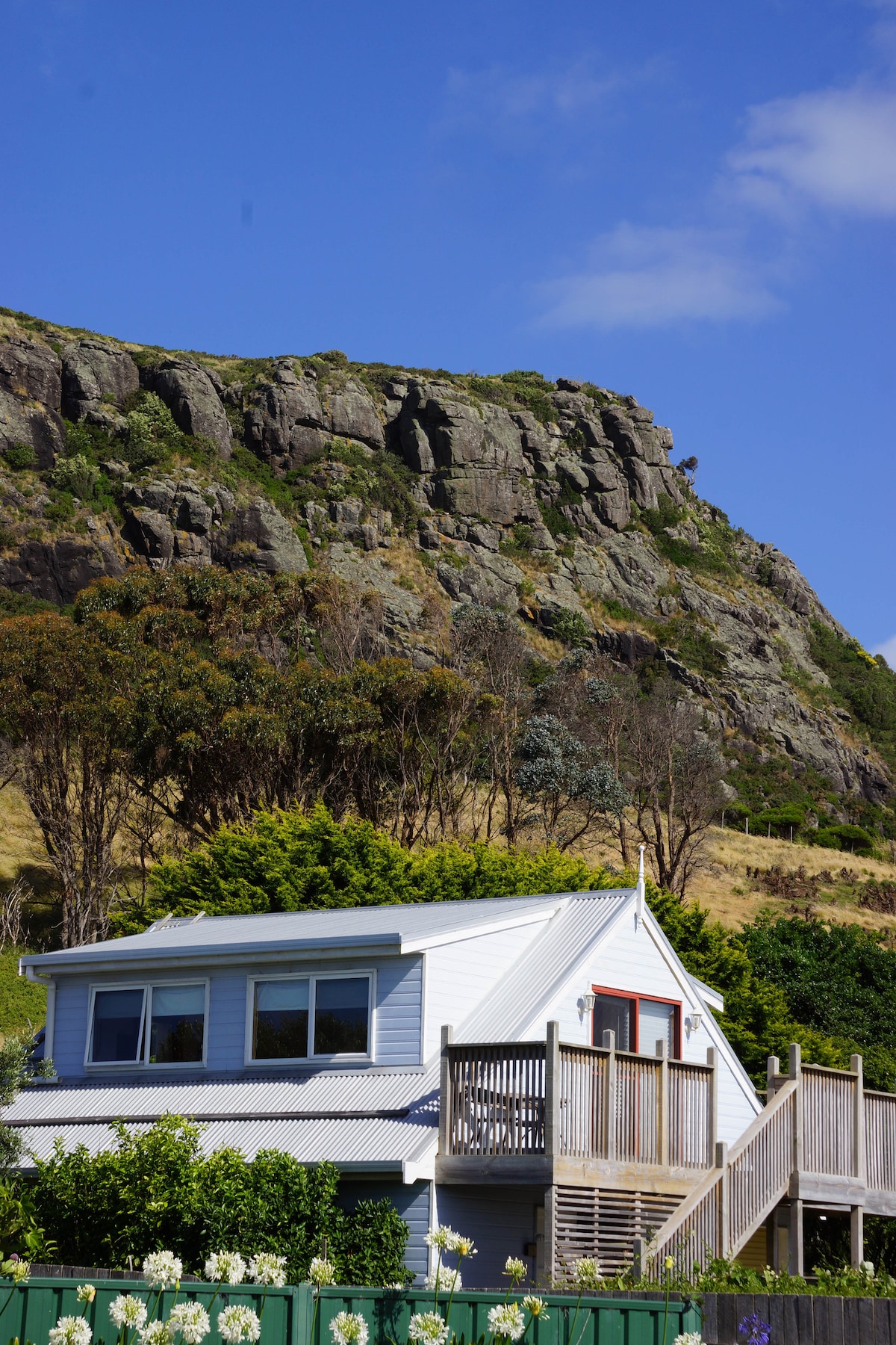 A two-story cottage is positioned against a backdrop of rugged cliffs and vibrant greenery. The bright exterior features a spacious deck with stairs leading up to the entrance. Lush vegetation and colorful flowers surround the property, enhancing the inviting setting.