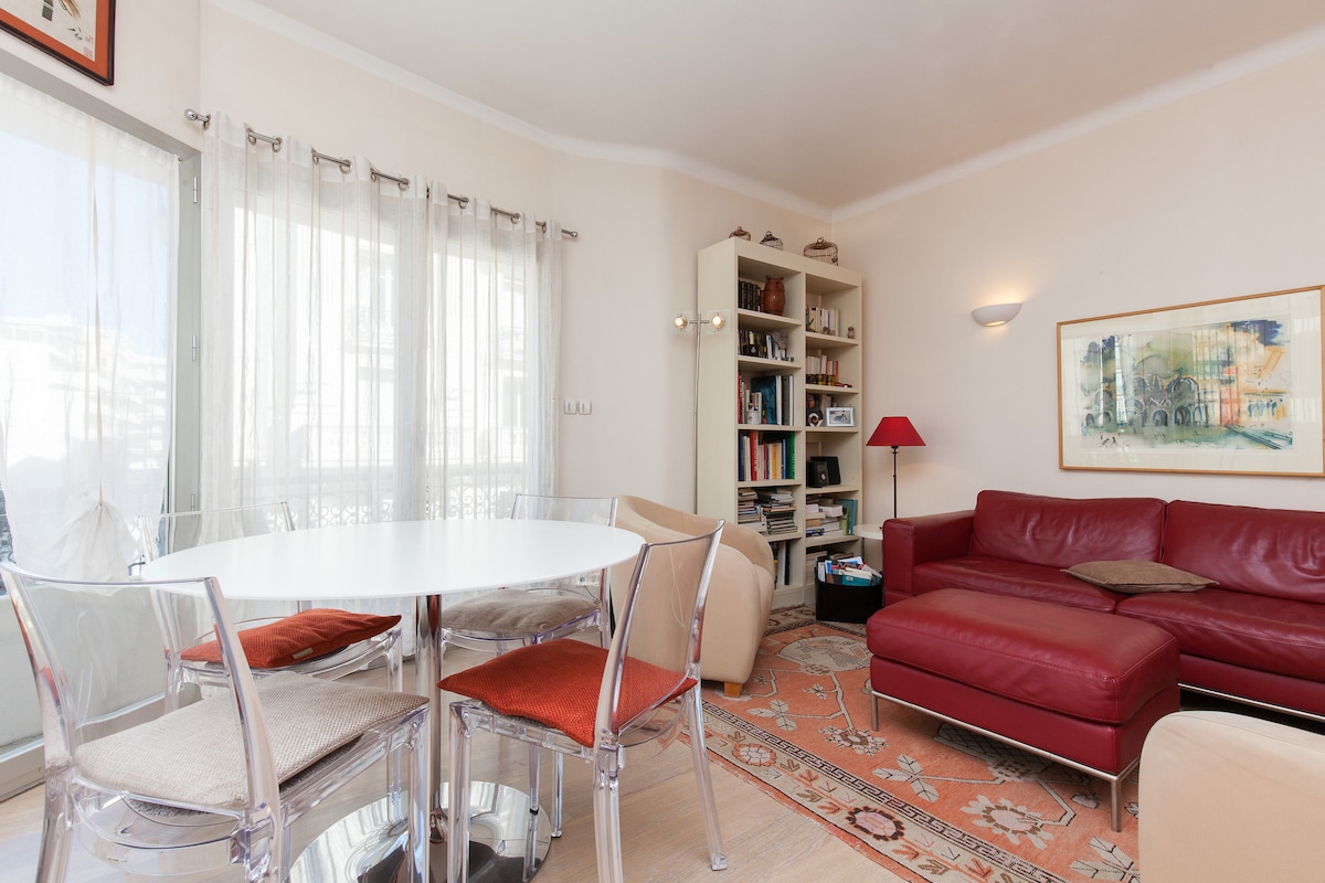 The living area features a round dining table with clear chairs and four cushions. A red leather sectional sofa is positioned against the wall, accompanied by a tall bookshelf containing various books. Natural light enters through large windows with sheer curtains.