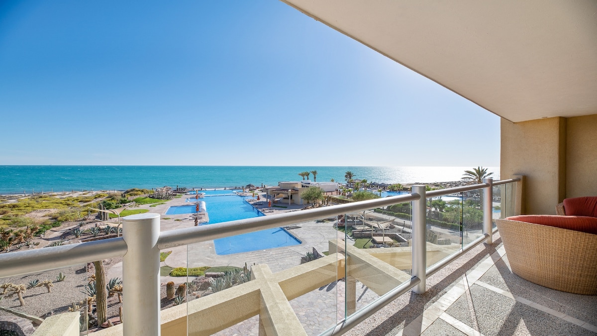 A spacious terrace overlooks a vibrant blue ocean and a beautifully designed swimming pool area. The horizon is clear and bright, showcasing a sunny day, while lounge seating provides an inviting place to take in the panoramic views.