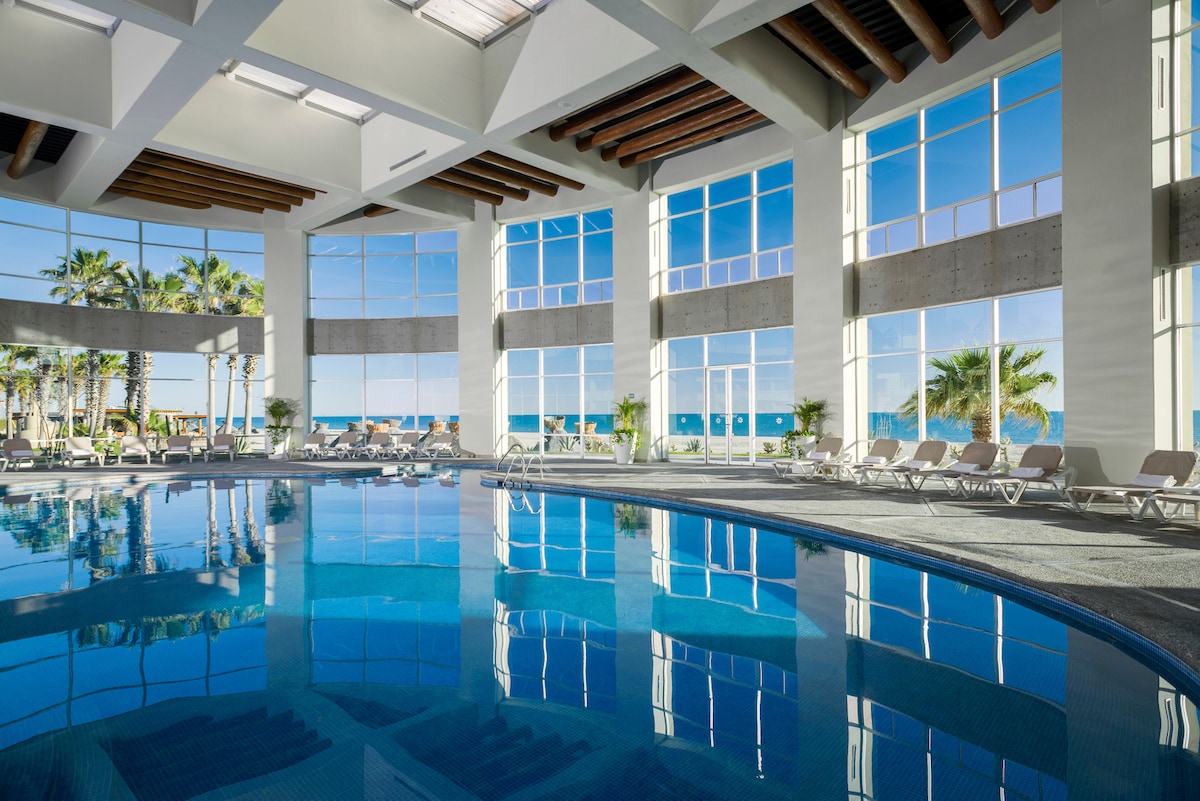 An indoor swimming pool is framed by large windows allowing natural light and views of palm trees outside. Comfortable lounge chairs are arranged around the water's edge, reflecting the serene blue tones of the pool, creating a tranquil atmosphere.